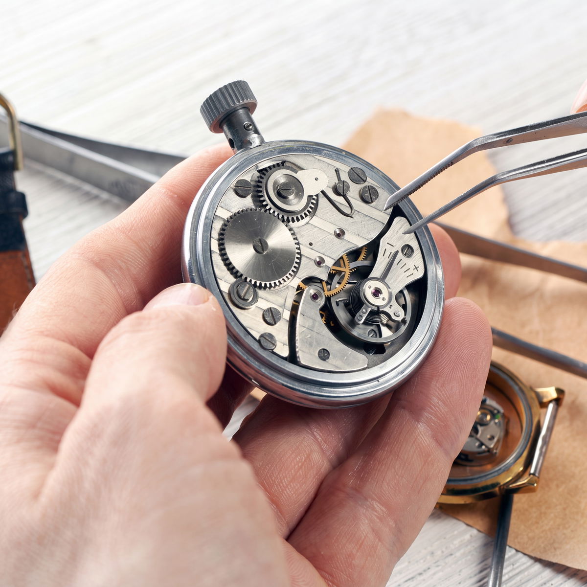 Watchmaker holding an open pocket watch movement and servicing it with tweezers at the bench