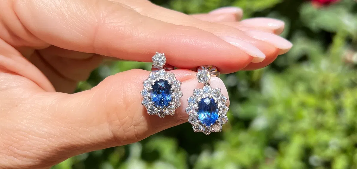 Oval sapphire and diamond cluster earrings held in hand against garden foliage