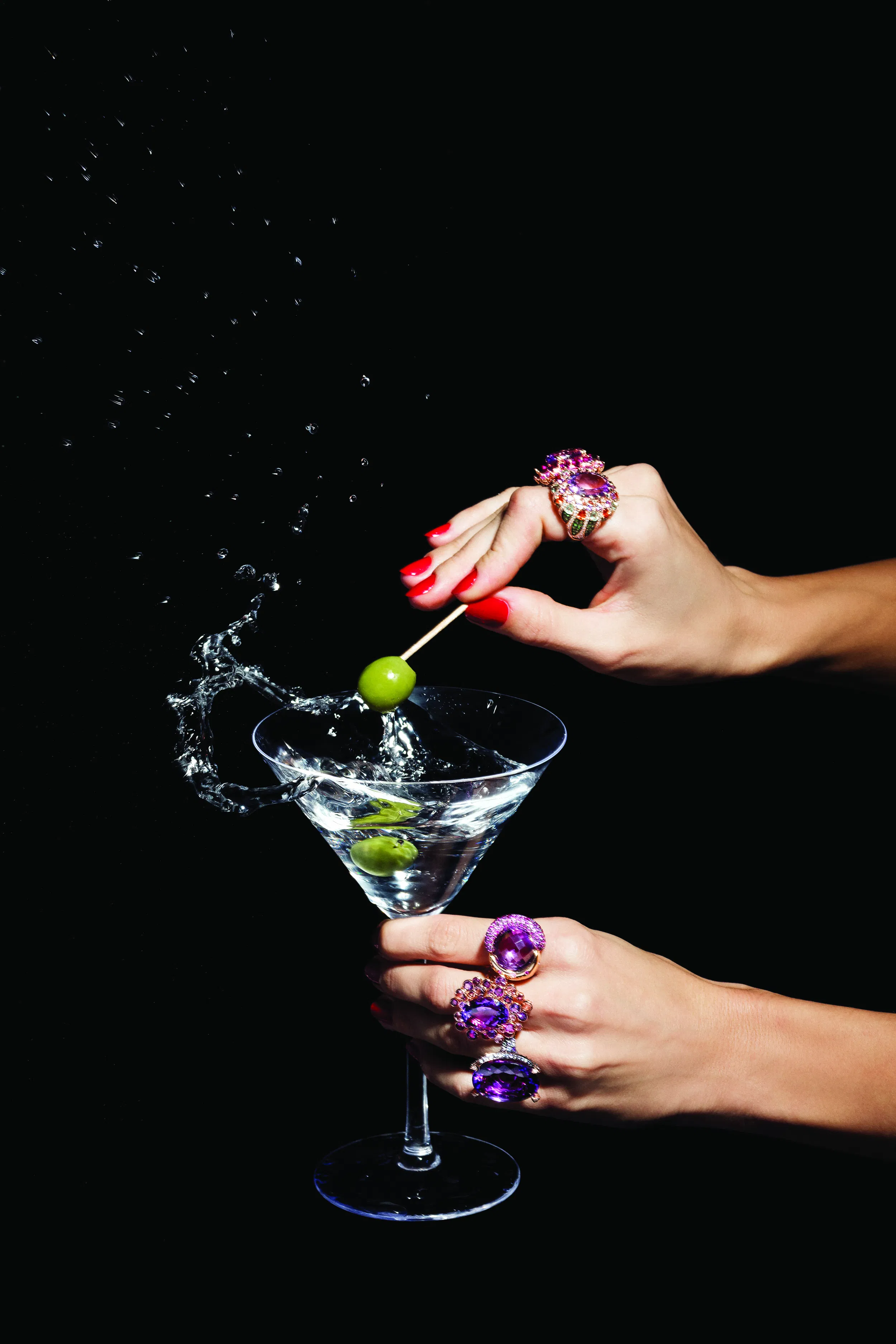 Model wearing statement gemstone rings garnishing a martini glass with an olive splash, high-fashion editorial style on black background