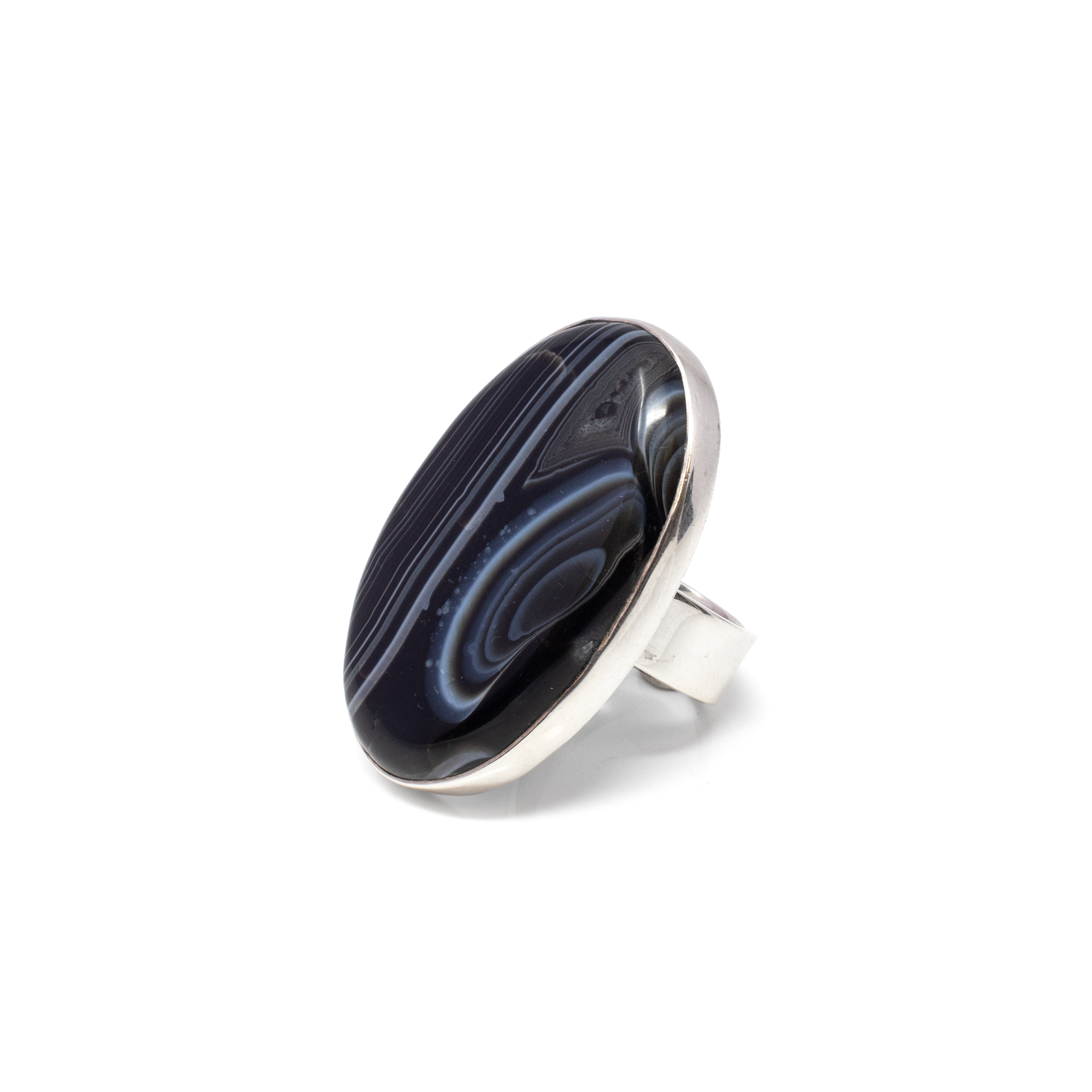 Elevate your style with the exquisite Banded Agate Ring, a stunning piece that marries nature's artistry with skilled craftsmanship. Featuring a captivating oval cabochon, this ring showcases the mesmerizing beauty of ebony agate, accented with intriguing coffee and white banding.Each ring displays a one-of-a-kind banded agate stone, renowned for its striking patterns and rich color palette. The ebony base, accented with coffee tones and delicate white striations, creates a visual symphony that’s both bold and elegant. Banded Agate Ring - Oval Cabochon With Silver Bezel - Ebony With Coffee & White Banding Szadj