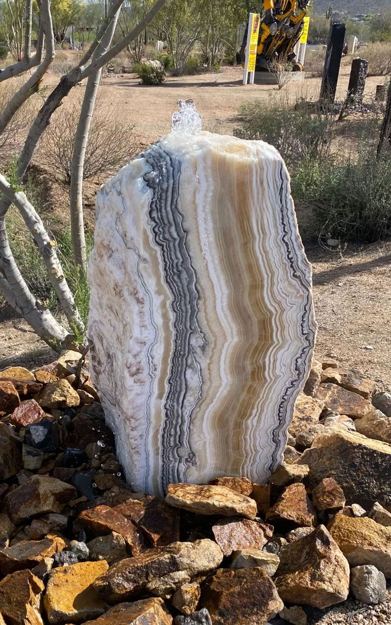 Elevate your outdoor sanctuary with the Zebra Onyx Fountain—an extraordinary work of natural art carved from authentic onyx. A single precision-cut face reveals the stone’s stunning banded layers in rich black and soft ivory, creating bold visual contrast and organic elegance. No two are alike, with each boulder selected for its dramatic striations and sculptural presence.This fountain is designed for custom placement and arrives without a basin, allowing flexible integration into your landscape, patio, or water feature. Whether as a tranquil focal point or a refined accent, the Zebra Onyx Fountain infuses any space with timeless beauty and the soothing presence of flowing water. IMPORTANT: the purchase price listed is for the FOUNTAIN ONLY and DOES NOT INCLUDE the cost of a basin, pump, parts or installation. Before purchasing a fountain online, we STRONGLY recommend you call us at (480) 575-4360 for a FREE consultation to discuss these additional costs and schedule a pre-site inspection. Also, free shipping does NOT apply to fountains, please call for a quote. Zebra Onyx Fountain ***NO Basin - Boulder with Single Side Cut & Striking Lines