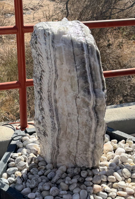 Introducing the Zebra Onyx Rustic Boulder Fountain – a stunning centerpiece that seamlessly combines natural beauty with sophisticated design. This exquisite water feature is expertly crafted from the finest Zebra Onyx, renowned for its unique veining that mirrors the striking appearance of a zebra's stripes. Let Rare Earth Gallery’s white-glove service make your fountain dreams come true—simply choose your fountain, tell us where it goes, and we’ll handle the rest, from delivery to installation. IMPORTANT: the purchase price listed is for the FOUNTAIN ONLY and DOES NOT INCLUDE the cost of a basin, pump, parts or installation. Before purchasing a fountain online, we STRONGLY recommend you call us at (480) 575-4360 for a FREE consultation to discuss these additional costs and schedule a pre-site inspection. Also, free shipping does NOT apply to fountains, please call for a quote. Zebra Onyx Rustic Boulder Fountain - 1 Cut Side
