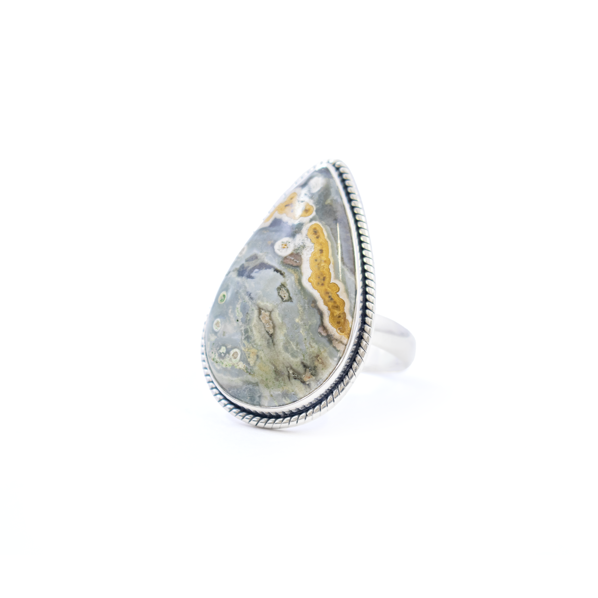 Embrace the enchanting allure of the sea with our exquisite Ocean Jasper Ring. This stunning piece features a mesmerizing pear-shaped cabochon of natural Ocean Jasper, renowned for its unique and vibrant patterns reminiscent of oceanic beauty. Each stone is a masterpiece of nature, promising a one-of-a-kind adornment that speaks volumes of elegance and sophistication.Set in high-quality 925 sterling silver, this ring boasts a beautifully crafted bezel that securely holds the Ocean Jasper in place, showcasing its captivating colors. The bezel is adorned with an intricate rope stamped edge, adding a touch of timeless charm and enhancing the ring’s classic appeal. Ocean Jasper Ring - Pear Cabochon Set in 925 Sterling Silver Bezel with Rope Stamped Edge sz10