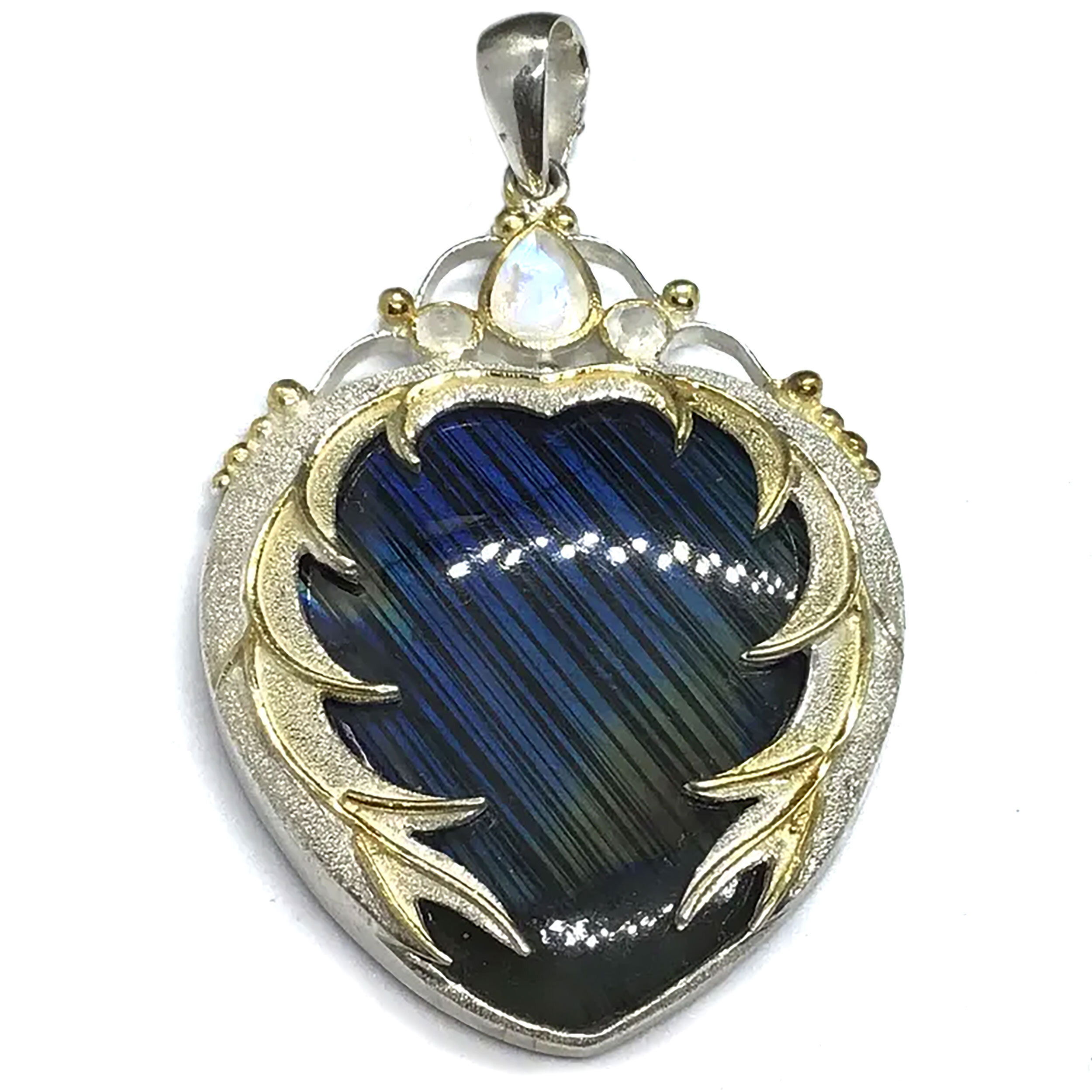The Labradorite Pendant is an enchanting composition of light and shadow, featuring a luminous blue labradorite cabochon alive with golden flash, bezel set in polished 925 sterling silver. Its heart-shaped sandblasted setting adds a subtle matte contrast, while the delicate gold plating frames the stone with warmth and elegance, evoking the feel of antique romance and modern sophistication.Above the central stone, two faceted moonstones shimmer with their own inner glow, lending a celestial balance to the piece. Graceful and ethereal, this pendant captures the magic of movement and light, making it a stunning accessory for both everyday elegance and special occasions. Ideal for those drawn to artisanal detail and the quiet power of natural gemstones, it’s a radiant celebration of beauty, craftsmanship, and inner glow. Labradorite Pendant - Blue with Yellow Flash Cabochon Bezel Set in 925 Sterling Silver Sandblasted Heart Setting with Gold Plating & Faceted Moonstones Above