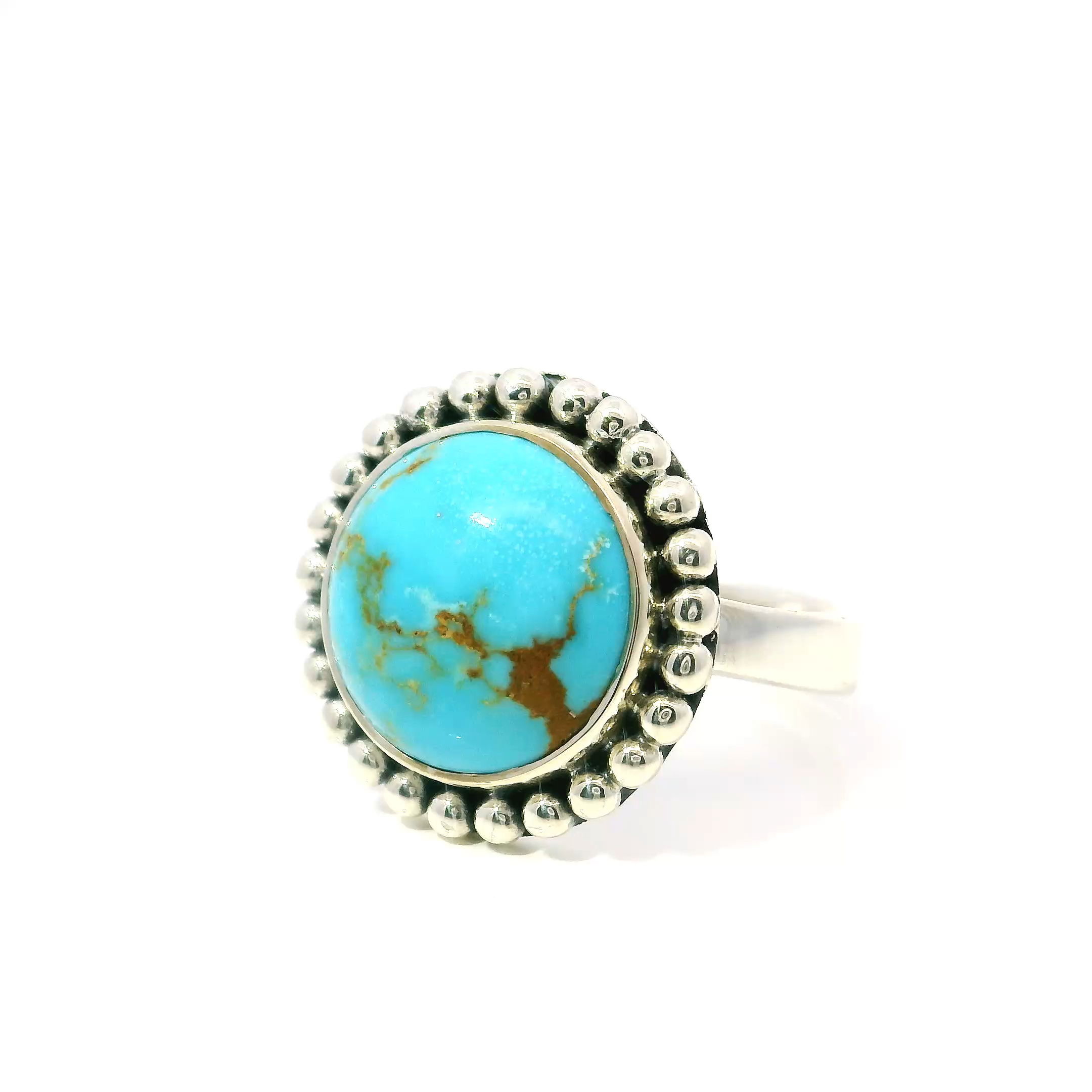 This New Mexican Turquoise Ring features a vibrant round cabochon with distinctive tan inclusions, each stone echoing the rugged beauty of the Southwest. Set in a 925 sterling silver bezel, the turquoise is framed by a delicate beaded edge that adds texture and refinement without overwhelming the stone’s natural character.Crafted in a comfortable size 7, this ring offers a timeless silhouette suited for daily wear or special occasions. The polished silver contrasts beautifully with the cool blue of the turquoise, resulting in a piece that feels both grounded and elegant. A quiet statement of heritage and artistry, it’s an ideal addition to any meaningful jewelry collection. New Mexican Turquoise Ring - Round Cabochon with Tan Inclusions Bezel Set with Beaded Edge in 925 Sterling Sivler sz7