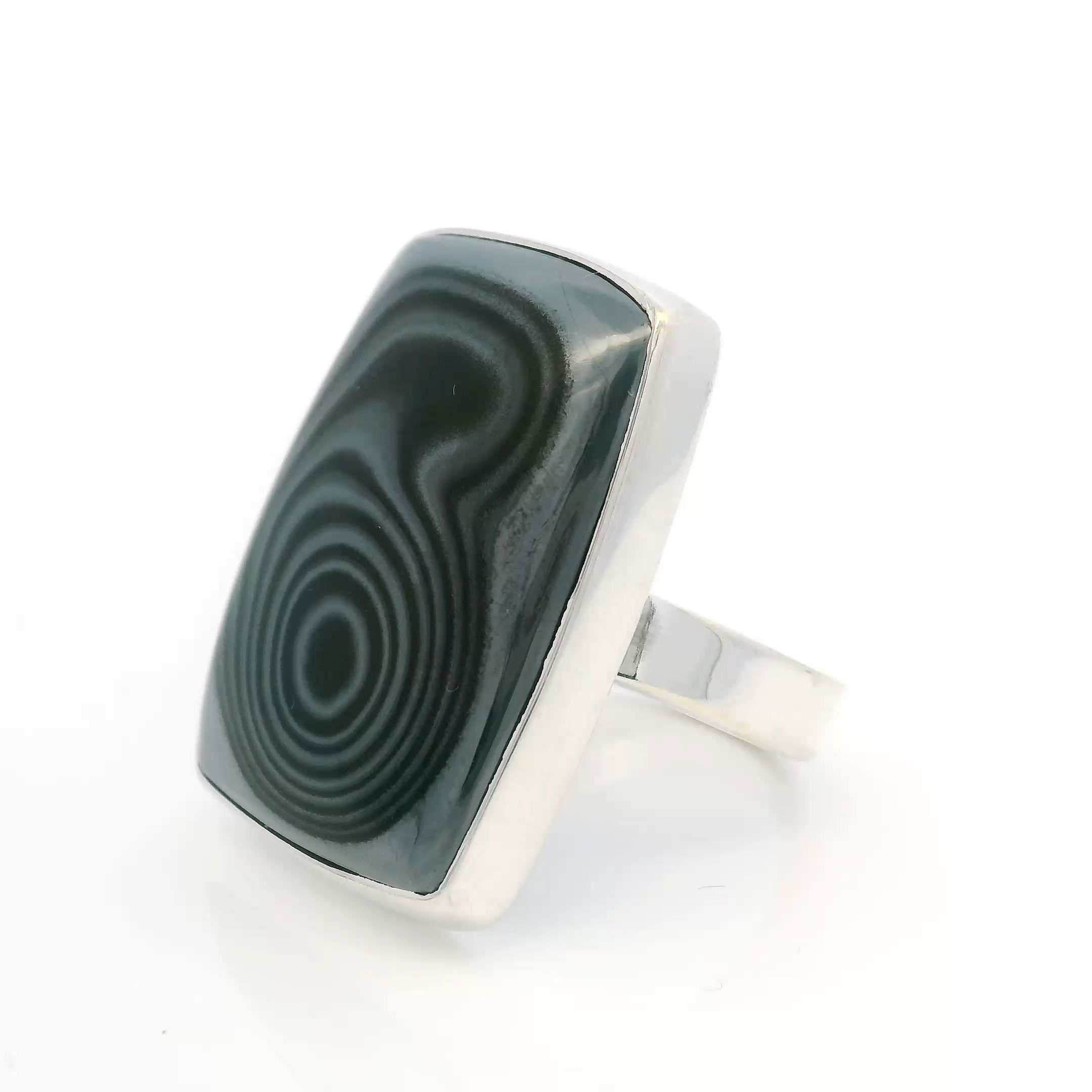 The Psilomelane Ring centers a rectangular cabochon alive with the mineral’s signature dark, fluid striations and a striking single bulls-eye—an arresting natural focal point shaped by geologic time. Set cleanly in a polished 925 sterling silver bezel, the stone’s dramatic presence is balanced by a minimalist frame that lets its patterns speak for themselves.With an adjustable band designed for comfort and versatility, this ring offers a refined yet grounded elegance. Perfect for women drawn to statement pieces rooted in the earth’s quiet mystery, the Psilomelane Ring brings bold sophistication to both everyday wear and special occasions. Psilomelane Ring - Rectangular Cabochon with Single Bulls-Eye Bezel Set in 925 Sterling Silver szAdj