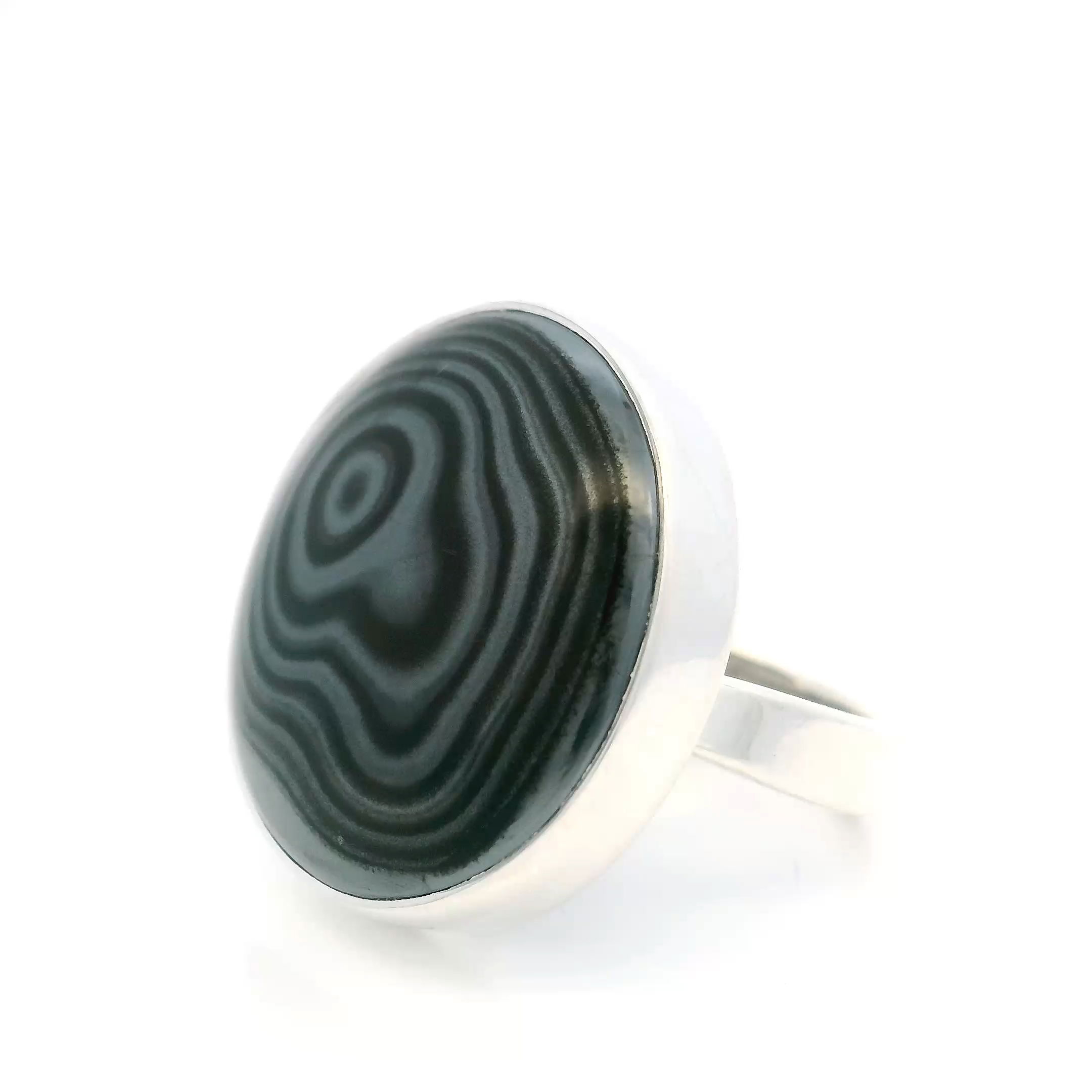 The Psilomelane Ring features a richly patterned round cabochon, its silvery-black surface alive with natural luster and storm-like swirls. Expertly bezel set in polished 925 sterling silver, the stone’s bold presence is matched by the simplicity and strength of its setting—timeless, grounded, and quietly powerful.With an adjustable band for a flexible fit, this piece merges natural wonder with everyday elegance. A perfect choice for those drawn to expressive stones and refined design, the Psilomelane Ring offers a distinctive accent that transitions effortlessly from day to evening. Psilomelane Ring - Round Cabochon Bezel Set in 925 Sterling Silver szAdj