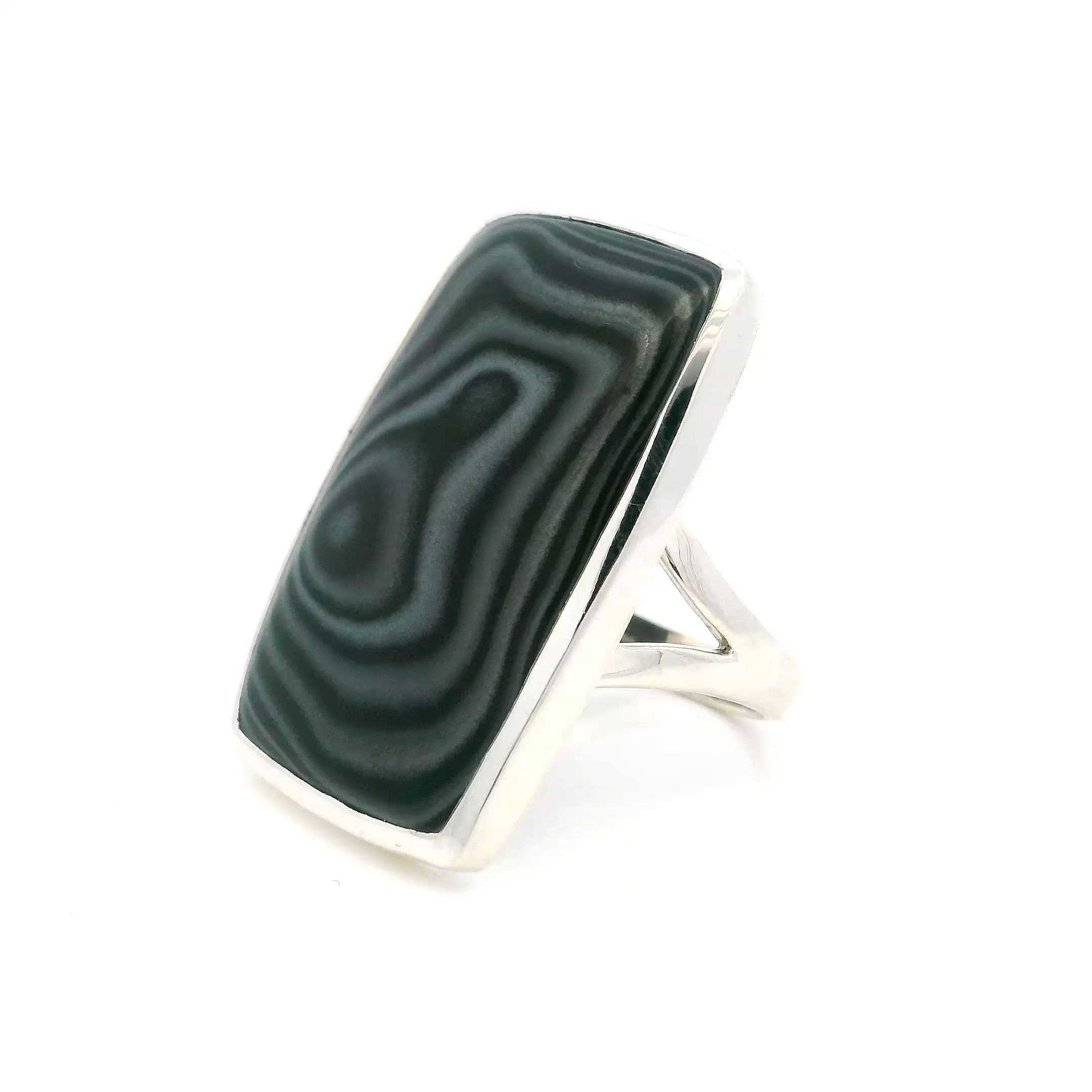 This striking Psilomelane Ring features a rounded rectangle cabochon with dark, silvery-black swirls that move like smoke across stone. The moody metallic patterns are bezel set in polished 925 sterling silver, grounding the piece in a clean, architectural silhouette that highlights the natural drama of the mineral.A wide, sculptural double band adds strength and comfort, balanced by a size 12 fit ideal for larger hands or those who favor bold, elemental jewelry. Refined yet raw, this is a powerful ring that carries the quiet sophistication of stone shaped by time. Psilomelane Ring - Rounded Rectangle Cabochon with Double Band Bezel Set in 925 Sterling Silver sz12