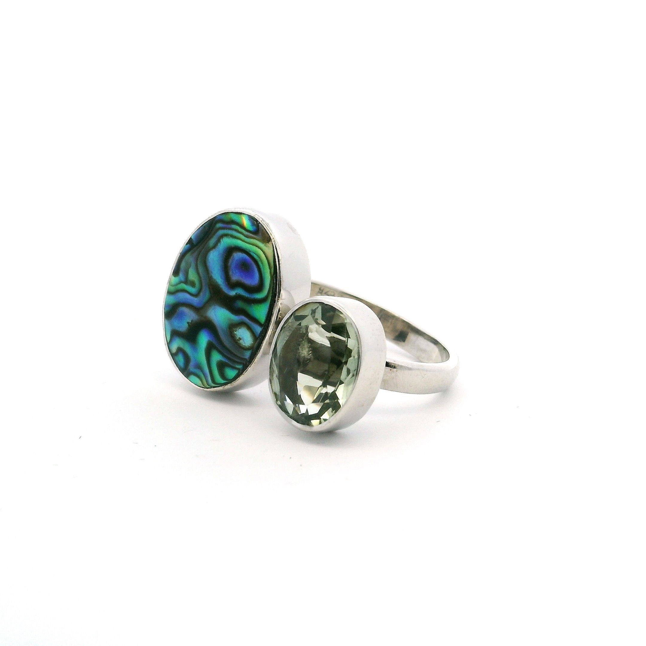 This Abalone Shell Ring with Oval Cabochon and Faceted Prasiolite in Adjustable 925 Sterling Silver captures the ocean’s iridescent beauty in a piece of wearable art. The oval cabochon of natural abalone shell shimmers with vibrant blues, greens, and purples—its surface echoing the shifting colors of the sea. Nestled beside it, a bezel-set faceted prasiolite adds a pale green sparkle, creating a striking interplay of tone and light.Set in a minimalist 925 sterling silver open band, this adjustable ring offers both comfort and a refined finish, allowing the stones to take center stage. A bold yet elegant accessory, it’s ideal for women seeking a ring that blends organic color with contemporary design. Abalone Shell Ring - Oval Cabochon with Faceted Prasiolite Oval Bezel Set on Open Band in Simple 925 Sterling Silver szAdj