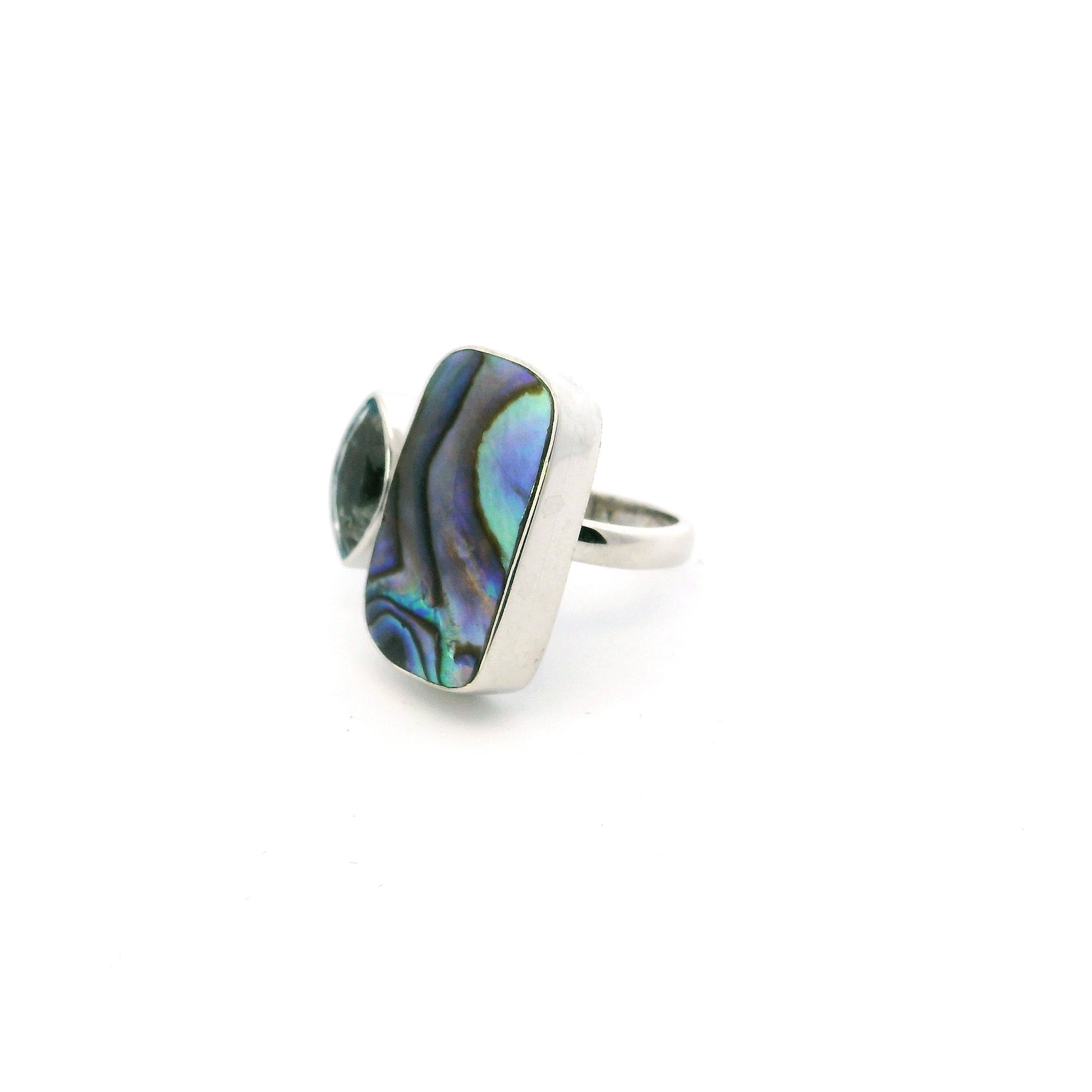This Abalone Shell Ring with Faceted Blue Topaz in Adjustable 925 Sterling Silver pairs oceanic iridescence with refined brilliance in a striking, modern silhouette. A rounded rectangular cabochon of natural abalone shell shimmers with vibrant tones of green, blue, and violet, its organic surface alive with color. Set beside it, a sharply faceted oval-cut blue topaz adds a cool, crystalline sparkle, bezel set for a clean, sculptural finish.The open band, crafted in polished 925 sterling silver, is fully adjustable for a comfortable fit and effortless styling. A bold yet graceful piece, this ring celebrates the union of natural textures and luminous gemstones, perfect for women drawn to beauty that feels both elemental and elegant. Abalone Shell Ring - Rounded Rectangular Cabochon with Faceted Blue Topaz Sharp Oval Bezel Set on Open Band in Simple 925 Sterling Silver szAdj