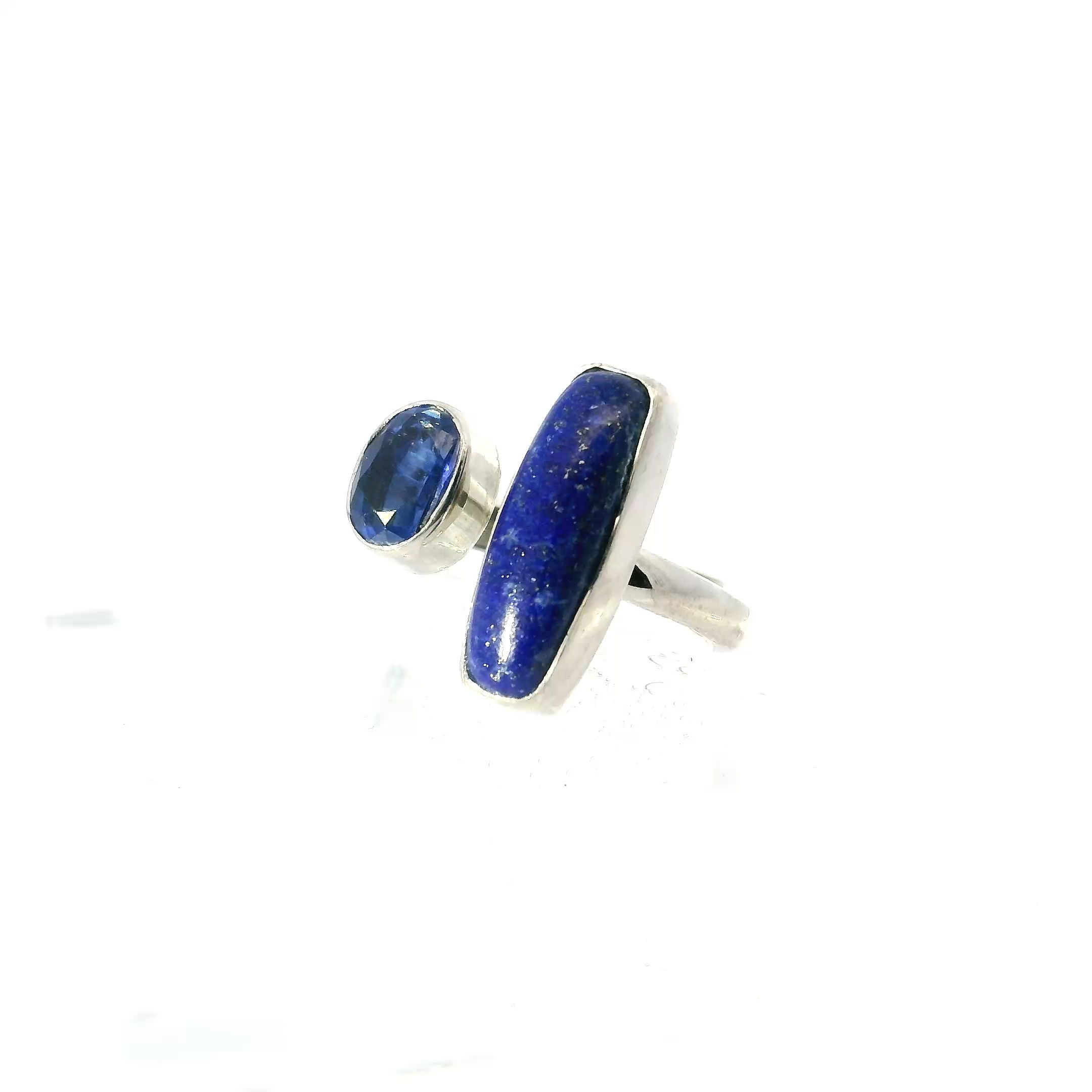 This Lapis Lazuli and Kyanite Ring with Narrow Rectangular Cabochon and Faceted Oval in 925 Sterling Silver features a slim, polished rectangle of deep blue lapis lazuli, delicately flecked with golden pyrite and bezel set for a sleek, modern finish. Beside it, a faceted oval blue kyanite adds shimmer and contrast, its east-west orientation lending a contemporary edge to the balanced design.Crafted in high-quality 925 sterling silver with an open band for easy adjustability, this ring combines comfort with artistry. Ideal for women seeking a refined, gemstone-forward piece with subtle flair, it’s perfect worn alone as a minimalist statement or stacked for added dimension. Lapis Lazuli Ring - Narrow Rectangular Cabochon with Faceted Kyanite Oval on Open Band Bezel Set in 925 Sterling Silver szAdj