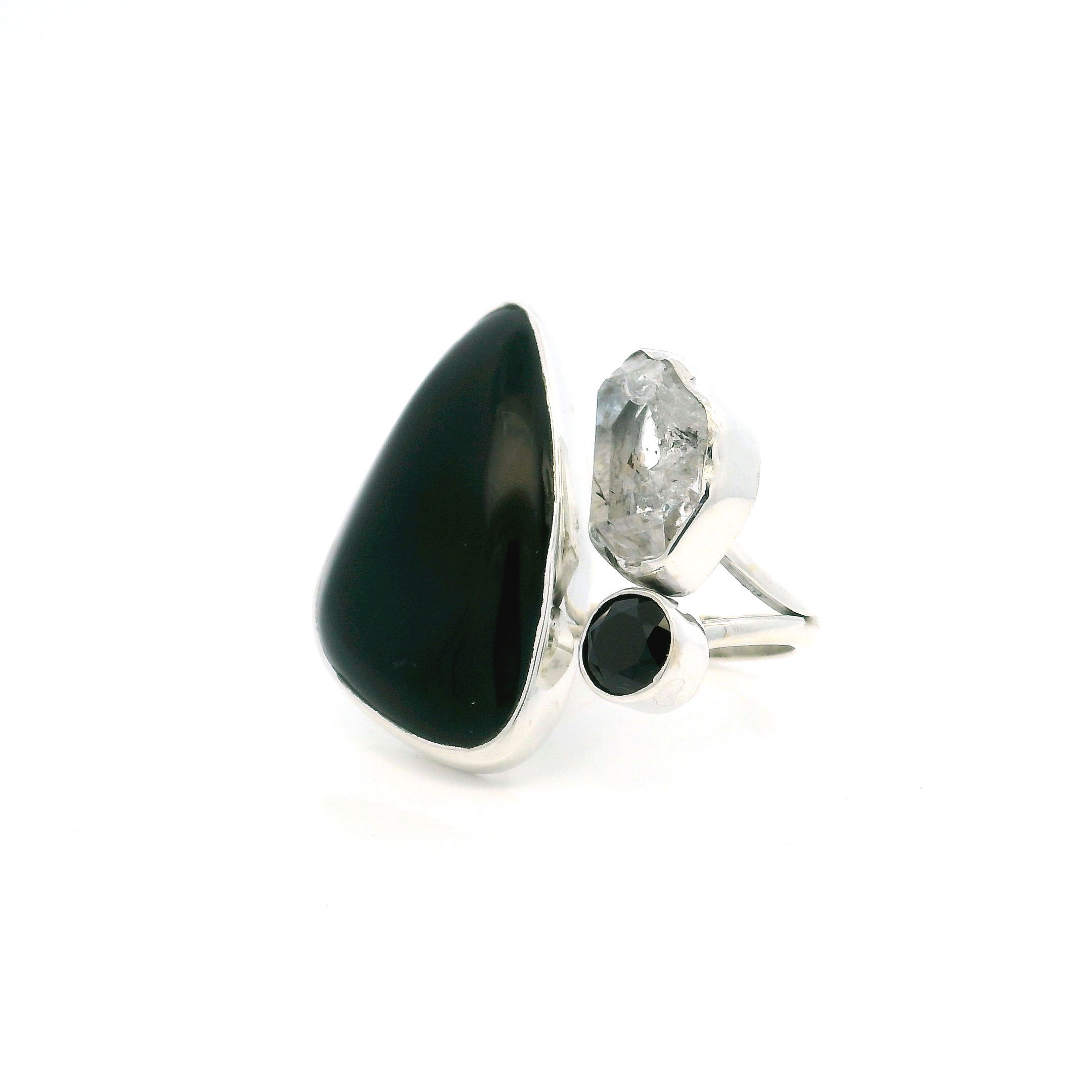 This Black Onyx Ring with Triangular Cabochon, Faceted Round Accent, and Herkimer Diamond in Adjustable 925 Sterling Silver is a bold yet refined statement piece that merges architectural lines with gemstone mystique. A sleek triangular cabochon of jet-black onyx anchors the design, its smooth surface exuding strength and serenity. Opposite it, a faceted round black onyx gleams with subtle fire, while a luminous Herkimer diamond adds unexpected brilliance—its raw clarity a striking contrast to the polished onyx forms.Crafted in high-quality sterling silver, the open band structure offers a sculptural, modern silhouette and allows for an adjustable fit. This ring effortlessly bridges the worlds of elegance and edge, perfect for women who embrace individuality and elevated design. Whether worn solo or layered, it’s a piece that commands attention with quiet intensity. Black Onyx Ring - Triangular Cabochon in Open Band with Herkimer Diamond & Faceted Black Onyx Round Bezel Set in 925 Sterling Silver szAdj