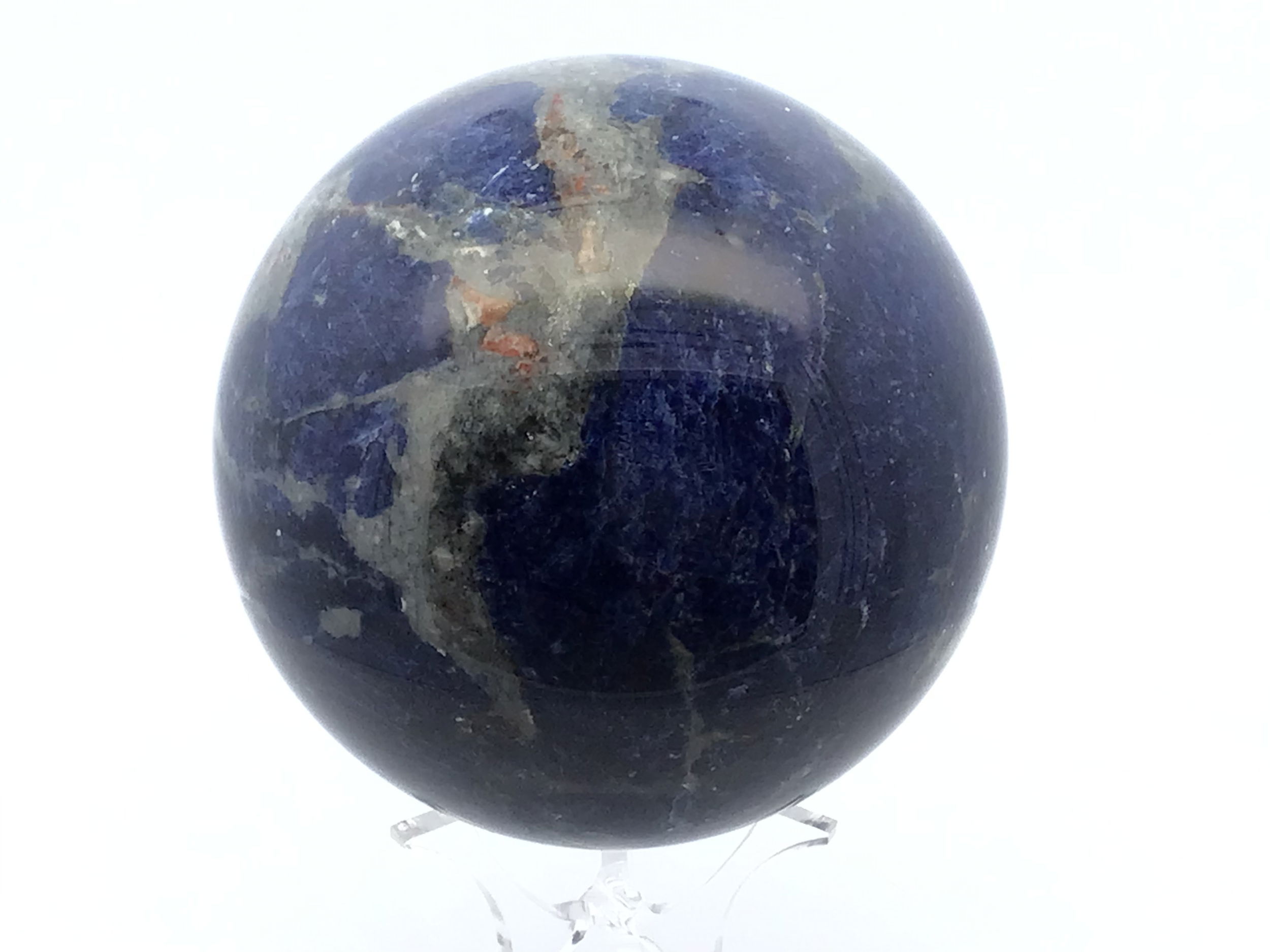 Hand-polished to reveal its striking interplay of deep blue and crisp white, this A+ Sodalite Sphere rests elegantly atop a clear lucite stand, blending organic beauty with modern design. Carefully selected for its rich coloration and minimal inclusions, each sphere radiates a calming presence, making it ideal for any décor, crystal grid, or meditation space.Sodalite is cherished for its ability to enhance intuition, promote mental clarity, and restore emotional balance. The sphere’s harmonious geometry further amplifies these properties, making it both a visual and energetic focal point. Whether displayed in a home office, living space, or sacred corner, this refined sodalite piece is a sophisticated symbol of peace and inner truth. A+ Sodalite Sphere with Lucite Stand