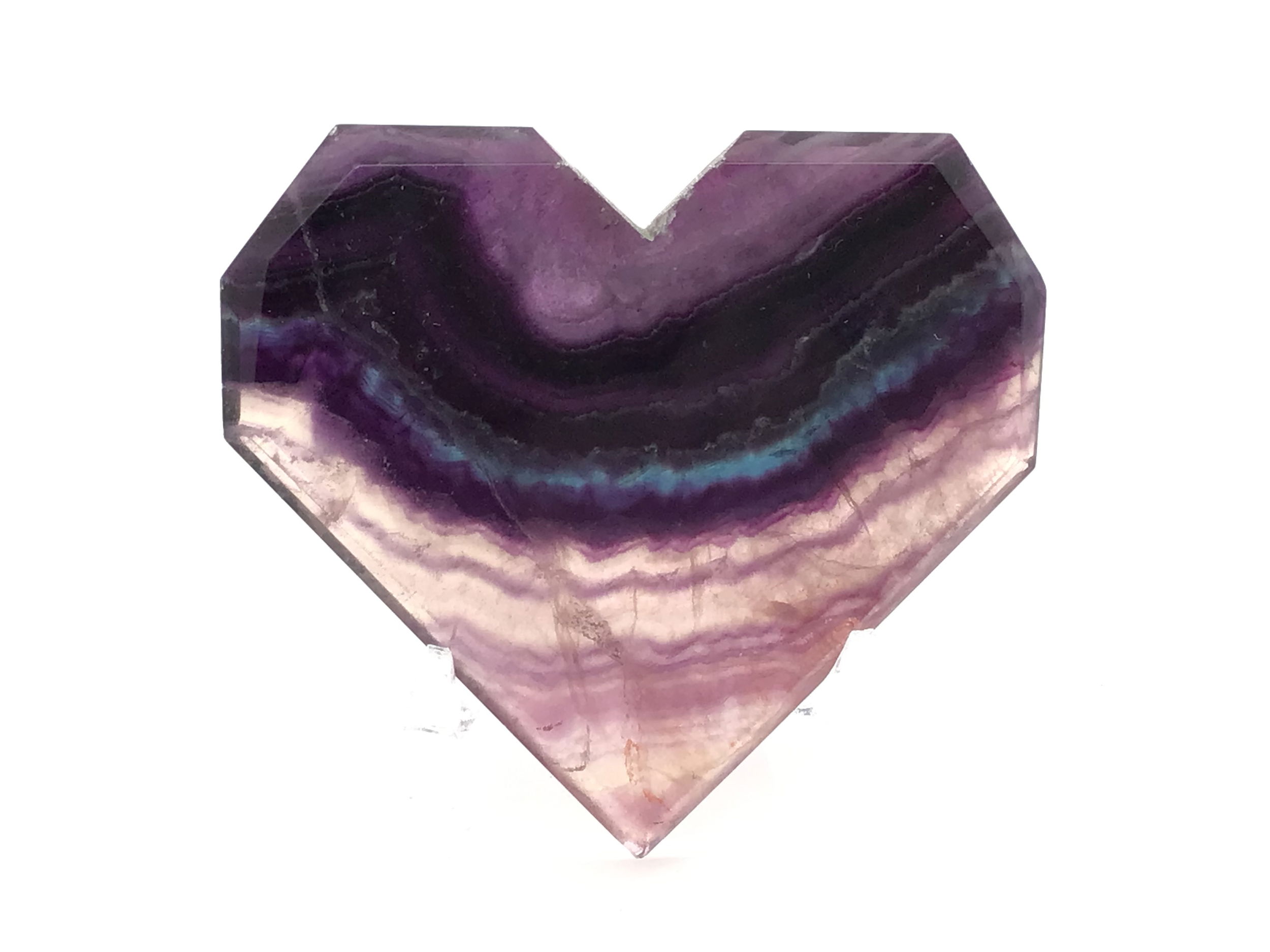 Illuminate your space with the enchanting beauty of the Rainbow Fluorite Heart – Flat with Faceted Faces on a sleek Lucite Stand. This captivating crystal showcases bands of tranquil blue accented by a whimsical, natural "smiling" stripe, making each piece truly unique. Expertly shaped into a geometric heart and adorned with delicately faceted faces, it catches and reflects light to reveal fluorite’s mesmerizing inner striations. Ideal as a centerpiece or an uplifting gift, this crystal heart beautifully harmonizes energy, promotes focus, and brings a touch of playful magic to any collection. Rainbow Fluorite Heart - Flat with Faceted Faces on Lucite Stand (Blue Smiling Stripe)