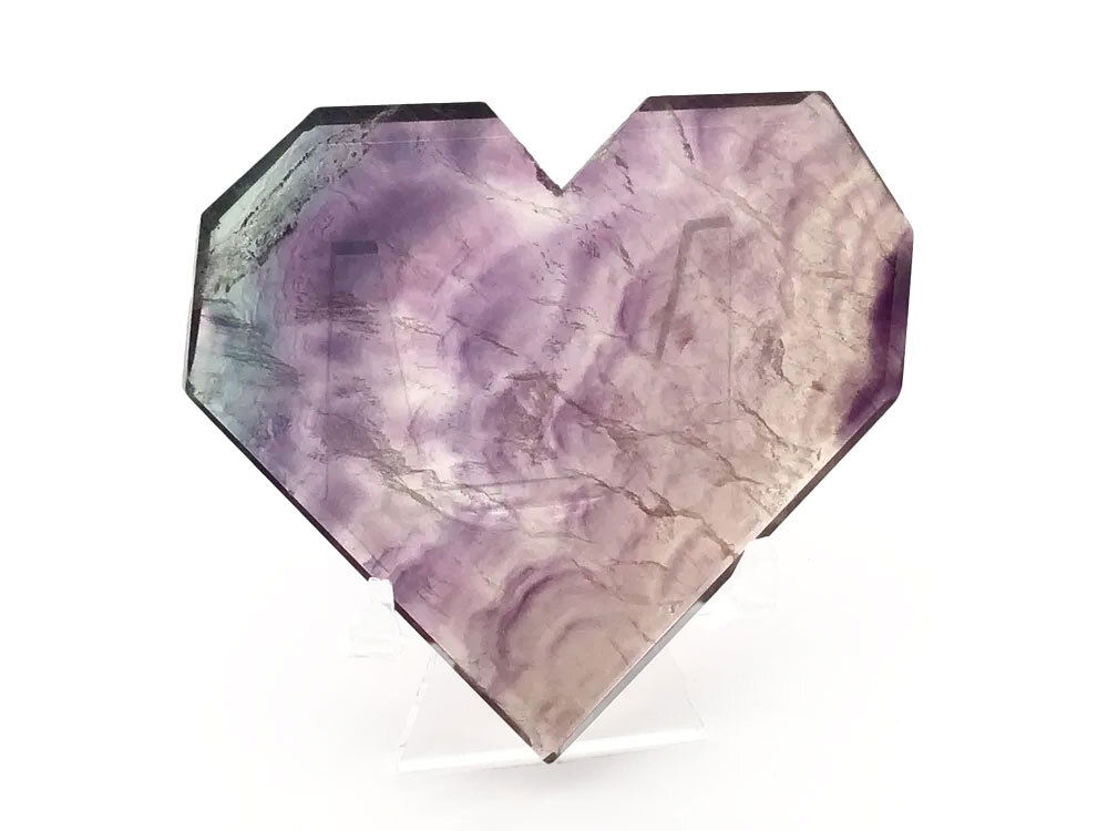 Elevate your space with the mesmerizing beauty of our Rainbow Fluorite Heart, expertly crafted to showcase both flat and faceted faces. Resting on a sleek lucite stand, this unique crystal heart features enchanting purple cloud formations swirling gracefully around a delicate green section—creating a captivating interplay of color and light. The geometric cut of the heart enhances the natural luminescence and inner clarity of high-quality fluorite, making it a striking centerpiece for any crystal collection, meditation area, or home décor arrangement. Rainbow Fluorite Heart - Flat with Faceted Faces on Lucite Stand (Purple Clouds with Small Green Section)