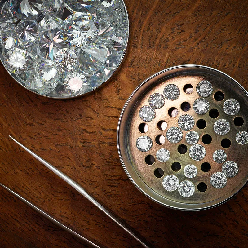 Top-down view of a wood-grain jeweler's workspace with a metallic sorting tray of small loose diamonds and fine tweezers