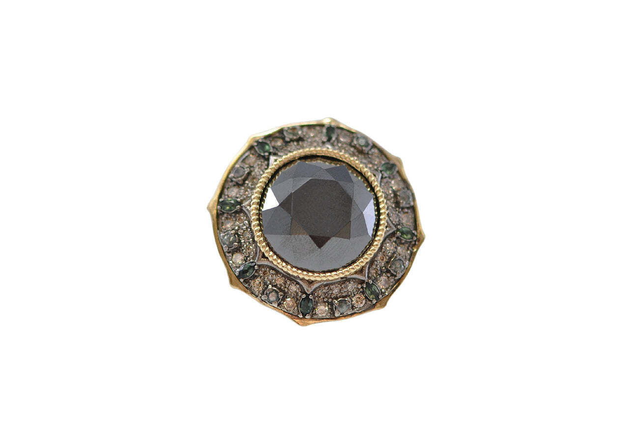 This stunning 18k yellow gold and blackened sterling silver cocktail ring is an absolute showstopper! Intricately designed with a textured round background, the 27mm pointed round ring features an eye-catching display of beautiful jewels. A 16mm hematite is surrounded by radiant blue grey sapphire, vibrant green tourmaline and soft champagne and white diamonds. Make a statement with this timeless and elegant piece that you'll cherish for years to come. Pointed Round Hematite Cocktail Ring